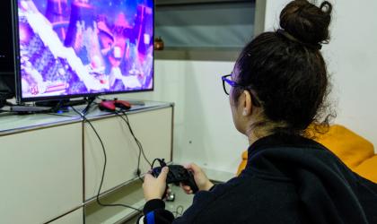 A droite, de dos est assise une jeune personne portant un chignon haut, des lunettes et un gilet noir. Elle tient à la main une manette connecté à un écran situé à gauche de la photo et sur lequel est affiché une partie de jeu vidéo.