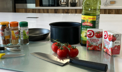 Sur un plan de travail blanc se trouve une planche à découper sur laquelle sont posés des couteaux, un économe, des tomates-cerises, deux boîte de coulis de tomate et trois flacons d'épices. Derrière tout cela sont posés une casserole, une poêle et une bouteille d'huile d'olive.