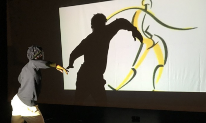 Accéder à l'événement "Spectacle : "Light Play" par Kivuko Compagnie". Visuel : Un danseur, de dos, dansant face à un écran diffusant une vidéo de figure dansante. L'ombre du danseur apparaît au gauche de celle de la figure à l'écran.