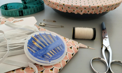 Accédez à l'événement "Atelier couture : raccommodage de vêtements". Visuel : Sur une table sont éparpillés une paire de ciseaux, un étui à aiguilles, un mètre-ruban, du tissu à motifs roses et des épingles à nourrice.