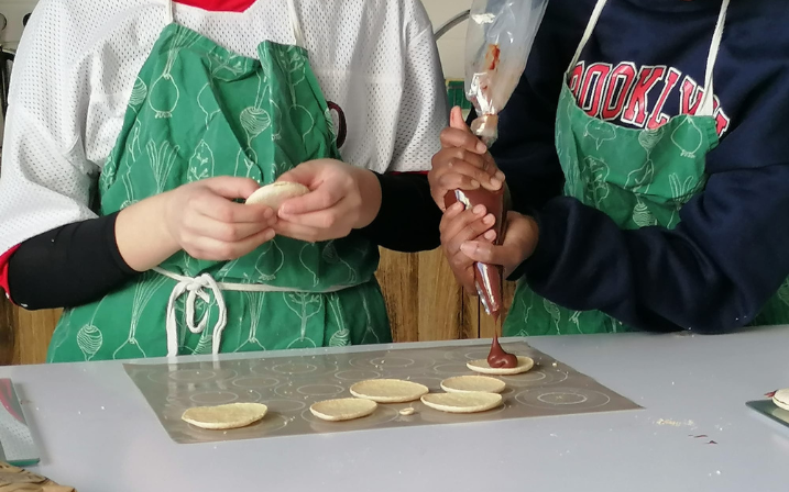 A gauche on voit une adolescente qui tient entre ses mains la croute d'un macaron tandis qu'à droite une autre adolescente presse une poche à douille remplie de chocolat sur des croutes de macarons posées sur un tapis de cuisson.